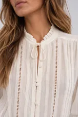Blusa de tejido texturizado color blanco, con cuello alto con volados y cordón para atar, cierre frontal de botones y mangas largas abullonadas con detalles de encaje y tablas verticales.