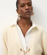 Cardigan de punto color crema con calce oversize, mangas amplias y cierre frontal mediante hebillas metálicas decorativas. Presenta una textura estructurada y puños ajustados.