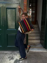 Cartera tipo tote de cuero con diseño de franjas verticales multicolores en tonos tierra, rojo y azul. Cuenta con correas trenzadas de cuero y cierre superior.