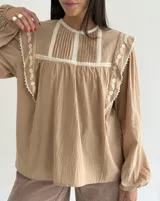 Blusa color beige de algodón con cuello redondo, mangas largas con puños elásticos y detalles de puntilla y bordados en el frente y hombros.