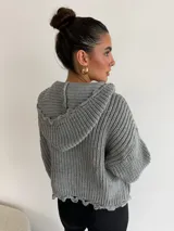 Buzo de punto grueso color gris, con cuello polo, cierre frontal parcial y capucha. Presenta un diseño de tejido acanalado con terminaciones desflecadas en el ruedo.