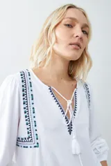 Remera blanca de manga larga con detalles bordados en tonos azules y verdes en el pecho y hombros. Presenta un escote en V con cordones ajustables terminados en borlas y puños elásticos.