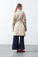 Trench de diseño clásico en color beige, con cierre frontal de botones, cinturón ajustable con hebilla a tono y mangas con trabillas en los puños.