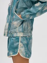 Short de estilo deportivo con pretina elástica, confeccionado en tela satinada con estampado tipo tie-dye en tonos azul y blanco. Presenta bordes con vivo en color crema y un pequeño logo bordado en la parte inferior.