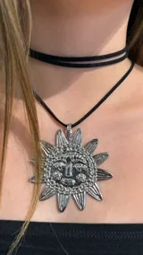 Gargantilla doble de cordón negro con un dije grande de metal plateado en forma de sol con rostro tallado.