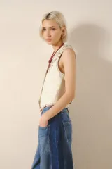 Chaleco de cuero de oveja color blanco hueso, de corte entallado, con cuello en V y cierre frontal de botones metálicos. Presenta bordados de tachas metálicas y piedras de colores en el frente, simulando motivos florales o de estrellas.