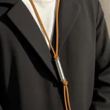 Collar largo con tira de gamuza marrón tostado y dije cilíndrico metálico plateado con detalles de pequeños cristales en los extremos.