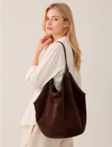 Cartera tipo hobo de gran capacidad confeccionada en símil gamuza color marrón. Presenta un diseño relajado y flexible con base reforzada y ribetes de cuerina a juego. Cuenta con una sola asa de hombro integrada a la estructura y un compartimento interior independiente.