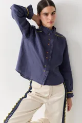 Camisa blanca de lino con cuello alto tipo gorguera, cierre frontal con botones de madera y detalles de ribetes en color mostaza en hombros y puños. Presenta volados en la botonera, cuello y puños.