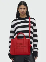 Bolso tipo tote de cuero negro, modelo The Leather Medium Tote Bag de Marc Jacobs. Presenta doble asa superior, correa ajustable para el hombro y el texto grabado "THE TOTE BAG MARC JACOBS" en el frente.