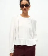 Blusa tipo bata de color blanco, confeccionada en viscosa, con cuello redondo, mangas largas con puños abotonados, detalles de entremedios verticales y un recorte con volado en el bajo.