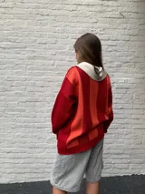 Sweater de punto rojo con rayas horizontales naranjas y blancas, cuello polo blanco y mangas largas. Presenta un diseño con letras curvas en el pecho.