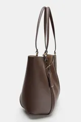 Bolso tipo shopper de hombro en color beige, con diseño estructurado, doble asa y detalle de pespuntes verticales en el cuerpo. Incluye un colgante decorativo tipo etiqueta y cierre imantado.
