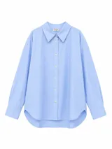 Camisa de corte oversize color azul claro, con cuello clásico, cierre frontal de botones y mangas largas con puños abotonados. Presenta un diseño con ruedo curvo.