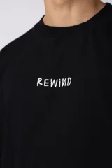 Remera negra de corte oversize confeccionada en tela pesada. Presenta un bordado frontal con la palabra 'REWIND' y un diseño de parche en la espalda con estética de papel roto.
