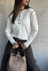 Remera de manga larga en color blanco, confeccionada en tejido acanalado. Presenta un diseño tipo henley con cuello redondo y una pequeña botonadura frontal, destacada por costuras a contratono en color oscuro que recorren los hombros y los puños.