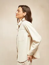 Camisa de manga larga color crudo, con cuello clásico, botones frontales y bolsillo de parche en el pecho. Presenta costuras decorativas en contraste.