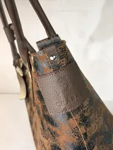 Cartera tipo tote de gran tamaño, confeccionada en cuero genuino con acabado texturizado y efecto desgastado en tonos tierra y negro. Cuenta con cierre superior metálico, interior forrado en cuero y un sujetador de asas con broche decorativo.