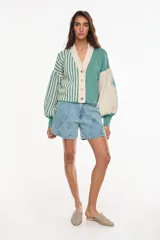 Cardigan tejido bicolor en tonos verde y crudo, con diseño de rayas verticales en la mitad derecha y liso en la izquierda. Presenta cuello en V, cierre frontal con botones y mangas abullonadas con puños ajustados.