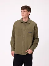 Camisa de gabardina color verde oliva, con cuello clásico, cierre frontal de botones y dos bolsillos frontales con parche. Incluye un pequeño parche con el logo de la marca en el bolsillo izquierdo.
