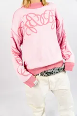 Sweater tejido reversible de corte boxy fit, con diseño gráfico de líneas curvas entrelazadas en el pecho y las mangas. Presenta un lado en tono rosa oscuro y el reverso en rosa claro, con cuello redondo y puños acanalados.