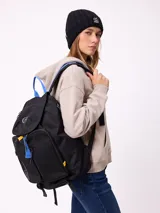 Mochila urbana marca Rusty de color negro, con múltiples compartimentos frontales, bolsillo de red lateral y detalles en azul y amarillo. Presenta el logo de la marca en la solapa superior y el texto 'Rusty Worldwide' en el frente.