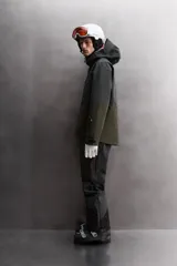 Cazadora deportiva de esquí con diseño bicolor en tonos negro y verde oliva. Presenta capucha amplia con visera, cierre frontal con cremallera termosellada, bolsillos frontales y en manga con cierres impermeables, y aberturas de ventilación en las axilas. Incluye faldón interior antinieve, puños ajustables y tecnología de rescate RECCO.