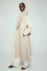 Chaleco largo de mezcla de lana color beige, de corte recto con manga sisa. Cuenta con cuello solapa con aplicación de pelo sintético extraíble, bolsillos laterales ocultos y un cinturón ajustable del mismo tejido para anudar en la cintura.