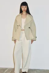 Trench corta de color beige con cuello de solapa y manga larga. Cuenta con bolsillos delanteros, cintura ajustable con cordones, bajo abullonado y cierre frontal cruzado con un botón visible. El interior está forrado con tela a cuadros.