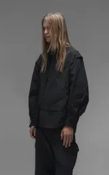 Chaqueta tipo overshirt de microfibra, color negro, con calce oversize y ruedo curvo. Presenta cierre frontal con broches a presión, bolsillos frontales de gran tamaño y cierres laterales a lo largo de las mangas para ajustar su amplitud.
