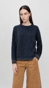 Sweater de lana color beige con calce holgado y cuello redondo. Presenta un bordado negro con diseño de arabescos en una de las mangas y detalles de rayas y puntos en contraste en el ruedo y los puños.