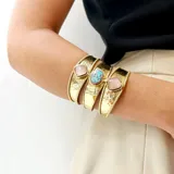 Brazalete ancho de bronce dorado, con textura martillada, compuesto por tres bandas unidas y adornado con tres piedras engarzadas: una central ovalada de nácar blanco y dos laterales cuadradas de nácar blanco.