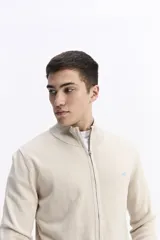 Campera tejida de algodón en color beige, con cierre frontal completo, cuello levemente elevado y puños y ruedo acanalados. Presenta un pequeño logo bordado en el pecho.