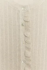 Chaqueta de punto canalé en color beige con hilo metalizado, cuello redondo, manga larga y cierre frontal con botones. Presenta detalles de volantes en la tapeta de los botones, el ruedo y los puños.