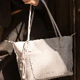 Cartera tipo tote de cuero marrón con costuras artesanales visibles, bolsillo frontal con el logo grabado, asas de hombro y detalles de flecos colgantes.