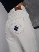 Pantalón vaquero de corte recto y tiro alto en color blanco marfil, con detalles de costuras en contraste y estampa decorativa en la parte inferior de las piernas, que incluye figuras de caballos y patrones geométricos. Presenta aberturas laterales en los tobillos.