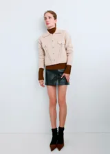 Chaqueta de punto color beige con cuello camisero, cierre frontal de botones metálicos y bolsillos en el pecho con solapa. Presenta detalles de tricotado texturizado y diseño corto.