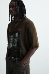Camiseta de punto de algodón con corte boxy fit, cuello redondo y manga corta. Presenta un estampado frontal con efecto desgastado en tonos blanco y rojo sobre un fondo marrón oscuro/verde oliva lavado.