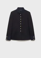 Chaqueta estructurada de corte recto en color negro, con cuello mao y cierre frontal de botones dorados con efecto joya. Presenta manga larga con puños abotonados y forro interior.