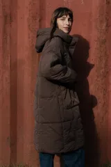 Campera larga acolchonada color marrón, con diseño midi, capucha amplia, cierre frontal con cremallera y broches, y bolsillos delanteros con tapa.