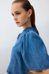 Blusa corta de denim liviano con mangas cortas abullonadas, cuello Peter Pan con volados y cierre frontal con tres tiras para atar.