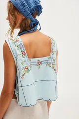 Top celeste sin mangas de algodón con escote cuadrado, bordado floral multicolor y líneas decorativas oscuras. Presenta un ruedo festoneado y laterales abiertos con tiras para ajustar.