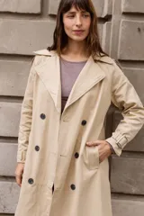 Trench clásico de largo midi en color beige, con diseño de doble abotonadura, solapas amplias, detalles en hombros y correas ajustables en los puños.