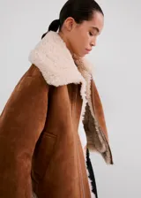 Chaqueta estilo aviador de ante sintético en color marrón con detalles de borrego sintético en el cuello, los puños, la solapa y el dobladillo inferior, con bolsillos laterales y cierre frontal con cremallera.