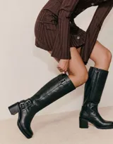 Bota de caña alta estilo biker, confeccionada en cuero negro. Presenta tres hebillas metálicas decorativas, cierre lateral y taco de madera.