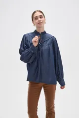 Blusa de lyocell en tono azul denim, con diseño holgado y frunces en el canesú. Presenta cuello alto con volados, cierre frontal mediante cordón ajustable y mangas largas con puños abotonados.