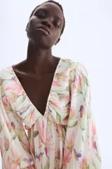 Vestido corto con textura plisada, estampado floral en tonos pastel (rosa, verde, amarillo, azul) sobre fondo crema. Presenta escote pronunciado en V con fruncido central y mangas globo largas ajustadas con lazos en los puños.