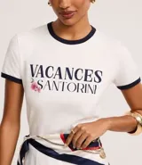 Blusa baby look de algodón color blanco, con cuello redondo y mangas cortas con ribetes azul marino. Presenta un estampado frontal con el texto "VACANCES SANTORINI" en azul marino y detalles florales en rosado.