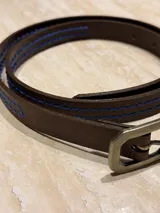 Cinturón fino de cuero marrón con costuras decorativas en hilo azul y hebilla metálica de diseño geométrico.