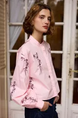 Camisa de algodón color rosa claro con cuello mao y cierre frontal de botones. Presenta un diseño bordado de flores en color violeta oscuro en la parte frontal y en las mangas largas con puños.